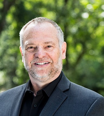 Photo of Christopher Weare smiling, wearing a suit and standing in front of greenery. 